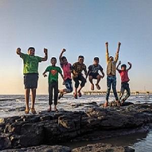 a group of children stood on rocks at a beach, jumping in the air with their arms up.