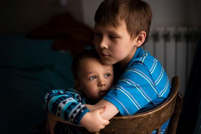 A young boy sat holding a baby with his arms protectively around the baby.